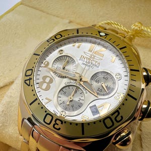 May include: Gold-toned Invicta chronograph watch with a white dial and three sub-dials. The watch features gold hands and markers, with the text "200 METERS (660FT)" visible. The watch is on a gold-toned bracelet.