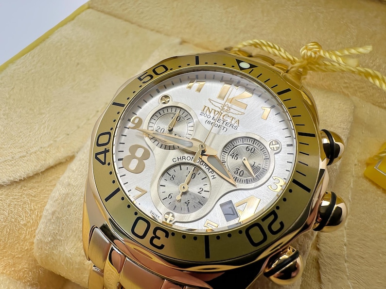 May include: Gold-toned Invicta chronograph watch with a white dial and three sub-dials. The watch features gold hands and markers, with the text "200 METERS (660FT)" visible. The watch is on a gold-toned bracelet.