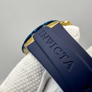 May include: Close-up of an Invicta watch. The watch features a navy blue band with the brand name embossed. The watch face has a blue bezel and gold accents. The watch is partially visible.