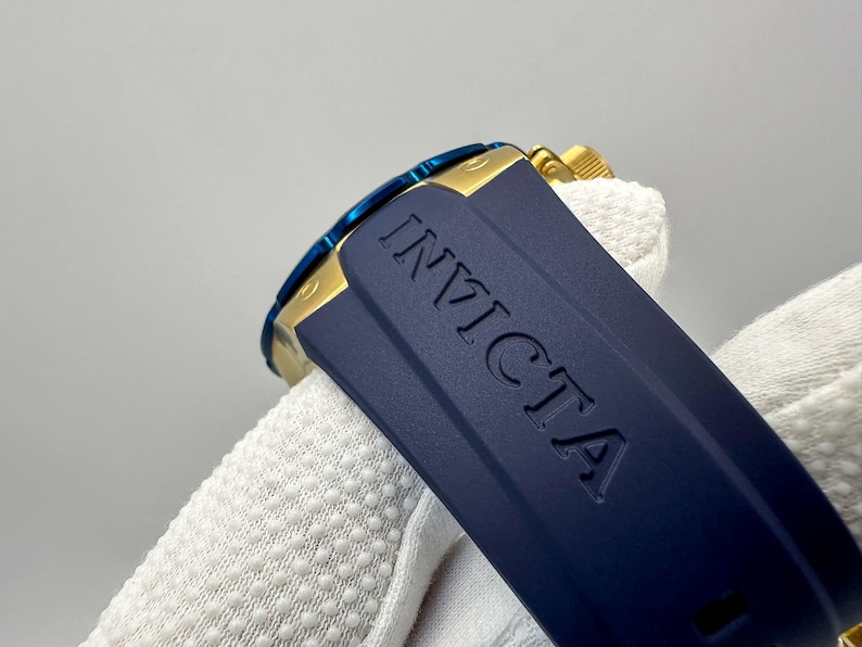May include: Close-up of an Invicta watch. The watch features a navy blue band with the brand name embossed. The watch face has a blue bezel and gold accents. The watch is partially visible.