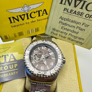 May include: A silver Invicta watch with a brown face, visible gears, and a stainless steel strap. The watch is presented in a yellow box with the Invicta logo and text. Accompanying documents are also visible.