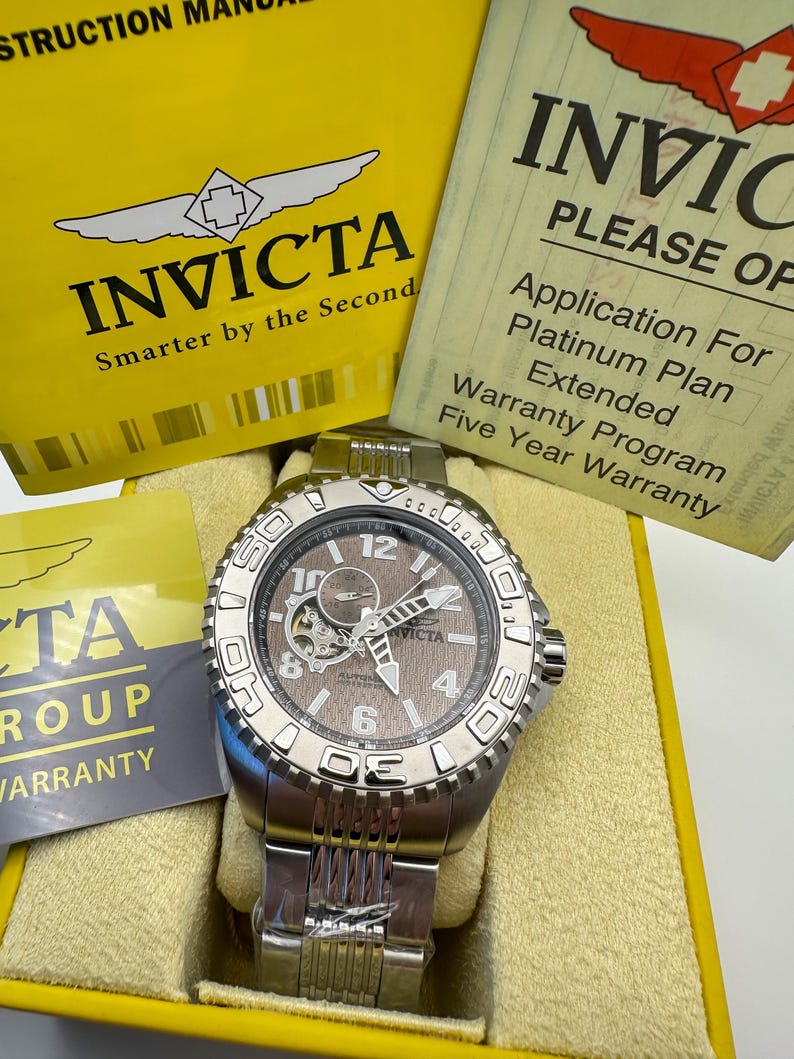 May include: A silver Invicta watch with a brown face, visible gears, and a stainless steel strap. The watch is presented in a yellow box with the Invicta logo and text. Accompanying documents are also visible.