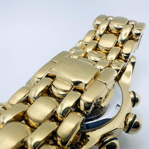 May include: Close-up of a gold-toned Invicta watch. The watch features a gold band with a clasp that has the Invicta logo. The watch face is partially visible, with a silver and black design. The watch has black accents.