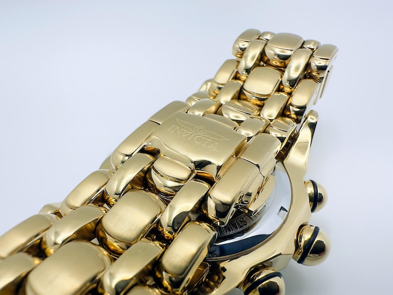 May include: Close-up of a gold-toned Invicta watch. The watch features a gold band with a clasp that has the Invicta logo. The watch face is partially visible, with a silver and black design. The watch has black accents.