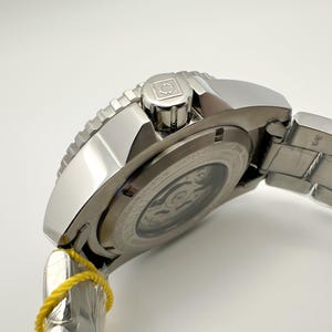 May include: A silver wristwatch with a stainless steel strap. The watch has a textured bezel and a crown with a logo. The back of the watch shows the inner workings. A yellow tag is attached to the strap.
