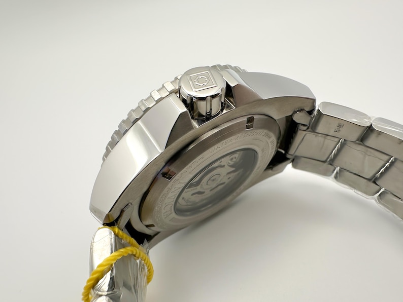 May include: A silver wristwatch with a stainless steel strap. The watch has a textured bezel and a crown with a logo. The back of the watch shows the inner workings. A yellow tag is attached to the strap.