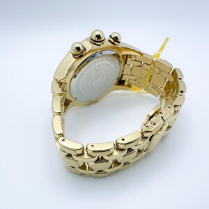 May include: A gold-toned wristwatch with a linked band and three small dials on the top. The watch has a round face with a silver-colored center. A yellow tag is attached to the band.