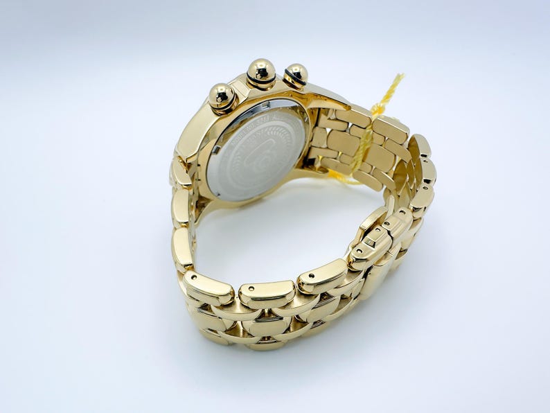May include: A gold-toned wristwatch with a linked band and three small dials on the top. The watch has a round face with a silver-colored center. A yellow tag is attached to the band.