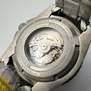 May include: A close-up of the back of a silver Invicta watch. The watch has a clear crystal back, revealing the inner workings of the automatic movement. Text includes "WATER RESISTANT 300M", "JAPAN AUTOMATIC MOVEMENT", and "STAINLESS STEEL".