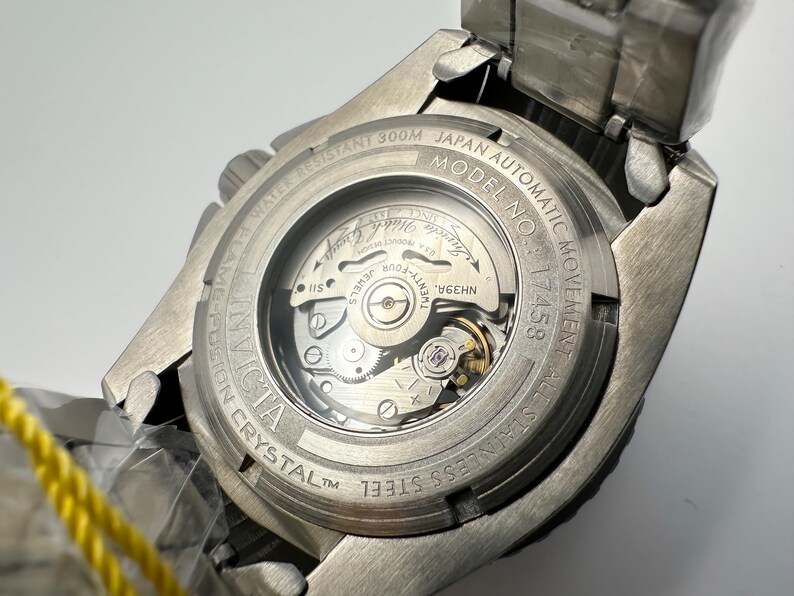 May include: A close-up of the back of a silver Invicta watch. The watch has a clear crystal back, revealing the inner workings of the automatic movement. Text includes "WATER RESISTANT 300M", "JAPAN AUTOMATIC MOVEMENT", and "STAINLESS STEEL".