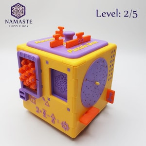 May include: A yellow and purple Namaste Puzzle Box with orange accents. The box has a variety of shapes and designs, including a flower, circles, and geometric shapes. The text "Namaste Puzzle Box" is visible on the top and side of the box. The level is 2/5.