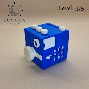 May include: A blue puzzle cube with white accents and knobs. The cube has the text "Level: 3/5" and the logo for Lunaria Puzzle Box. The cube has various buttons and dials.
