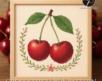 Puede incluir: Patrón de punto de cruz con dos cerezas rojas con hojas y tallos verdes, rodeadas por una corona floral. El diseño está enmarcado en un marco cuadrado de madera clara. El texto "CROSS STITCH PDF PATTERN" está en la parte inferior.