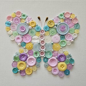 May include: A colorful butterfly art piece made from various pastel-colored buttons. The buttons are arranged on a white canvas, with pearl accents outlining the butterfly's wings. The butterfly has brown antennae.