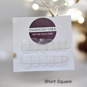 May include: Clear, short square press-on nails are displayed on a white card with the text "Enchanted Nails" and "Every nail tells a story!". The image is set against a blurred background with string lights and the words "Short Square".