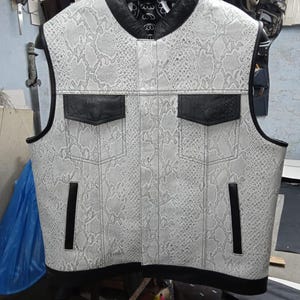 Puede incluir: Un chaleco sin mangas con estampado de piel de serpiente en tonos grises y blancos. El chaleco presenta detalles en cuero negro en el cuello, los bolsillos y los ribetes. Dos bolsillos son visibles en el pecho, con detalles verticales negros en los laterales.
