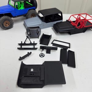Könnte beinhalten: Eine Sammlung von Miniatur-Modell-Jeep-Teilen in verschiedenen Farben, darunter Blau, Grau und Schwarz. Die Teile sind auf einer weißen Oberfläche angeordnet und zeigen die Komponenten eines kleinen Fahrzeugs, einschließlich Fahrgestell, Karosserie und Überrollkäfig.