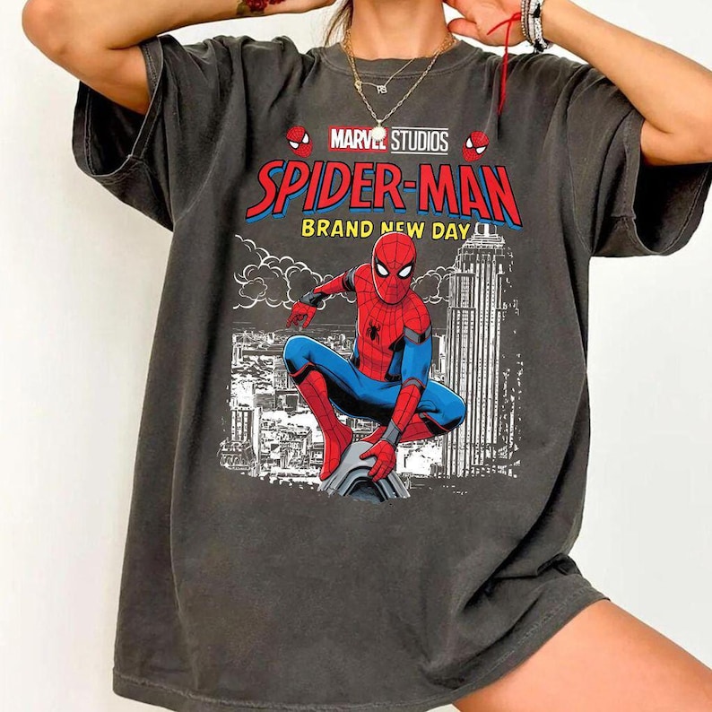 Może przedstawiać: Ciemnoszara koszulka z grafiką Spider-Mana z Marvel Studios. Projekt zawiera napisy "Spider-Man Brand New Day" oraz ilustrację Spider-Mana w czerwono-niebieskim kostiumie, na tle panoramy miasta.