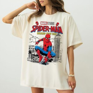 Może przedstawiać: Kremowa koszulka z grafiką Spider-Mana w pozycji kucznej, z napisem "MARVEL STUDIOS SPIDER-MAN BRAND NEW DAY". Projekt zawiera tło z krajobrazem miejskim i logo Marvel Studios.