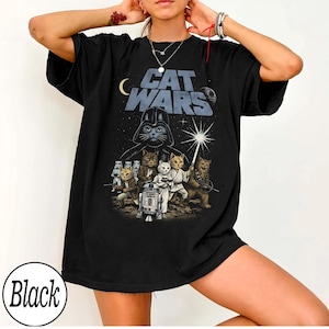 Może przedstawiać: Czarny t-shirt z grafiką "Cat Wars". Projekt zawiera kocią wersję Dartha Vadera, księżyc, gwiazdy i różne koty w kostiumach z motywem Gwiezdnych Wojen. Tekst "Cat Wars" jest wyświetlany nad centralnym obrazem.