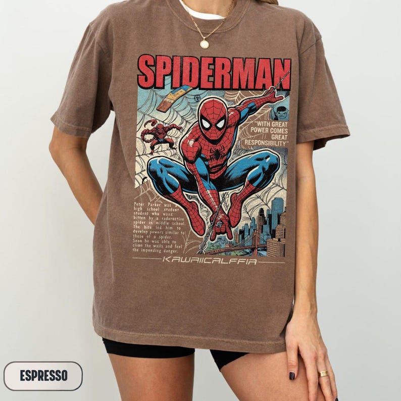May include: Espresso-colored t-shirt featuring a comic book-style illustration of Spider-Man. The design includes the word "SPIDERMAN" in red, blue, and white, along with text and a cityscape background. The shirt has short sleeves.
