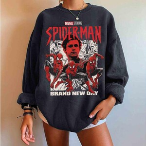 Puede incluir: Sudadera negra con un dise&ntilde;o de Spider-Man. El texto "SPIDER-MAN" es rojo y "BRAND NEW DAY" es blanco. El logotipo de Marvel Studios est&aacute; en la parte superior. El dise&ntilde;o incluye im&aacute;genes de Spider-Man en acci&oacute;n.
