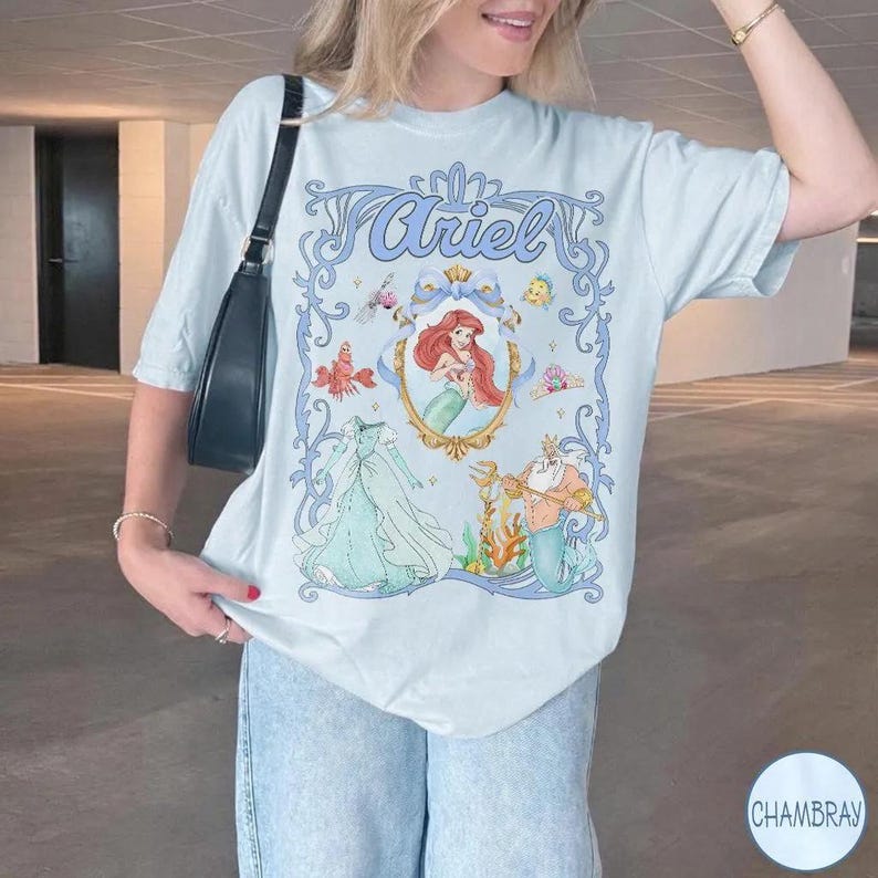 May include: Light blue t-shirt featuring a framed illustration of Ariel from The Little Mermaid. The design includes the name "Ariel" in script, a portrait of Ariel, and other character illustrations. The shirt is a relaxed fit.
