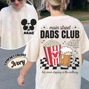 Pode incluir: Camiseta cor marfim com um design do Mickey Mouse na frente e um gráfico "Main Street Dads Club" nas costas. O gráfico nas costas inclui pipocas, um caneco de cerveja e texto. A camisa é da marca Comfort Colors.