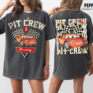 May include: Dark gray t-shirt with a front graphic of a red cartoon race car, checkered flags, and the text "PIT CREW Daddy". The back of the shirt features the same car and text, with additional text and flags.
