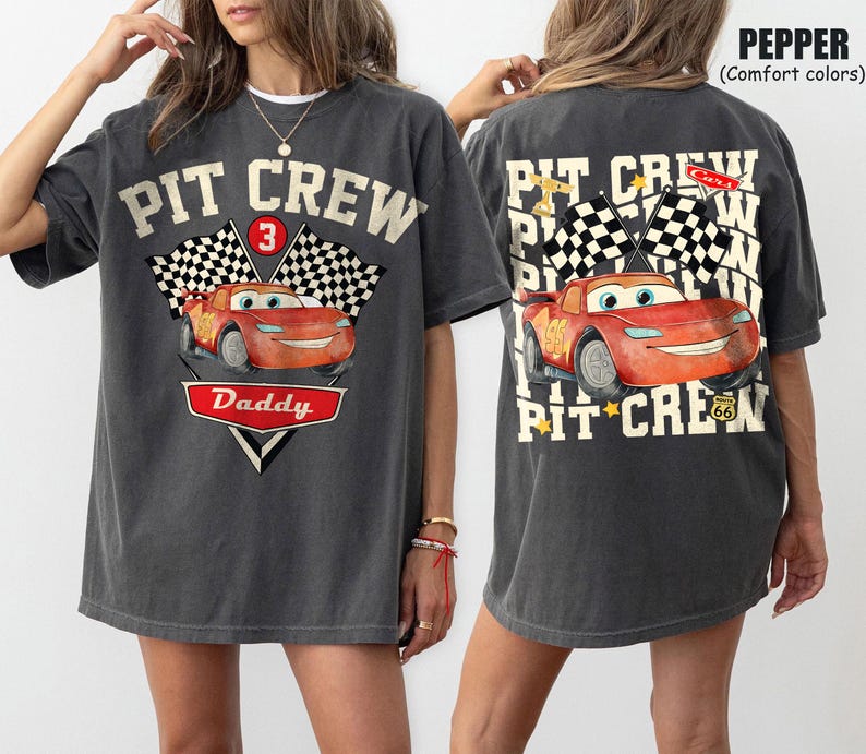 May include: Dark gray t-shirt with a front graphic of a red cartoon race car, checkered flags, and the text "PIT CREW Daddy". The back of the shirt features the same car and text, with additional text and flags.