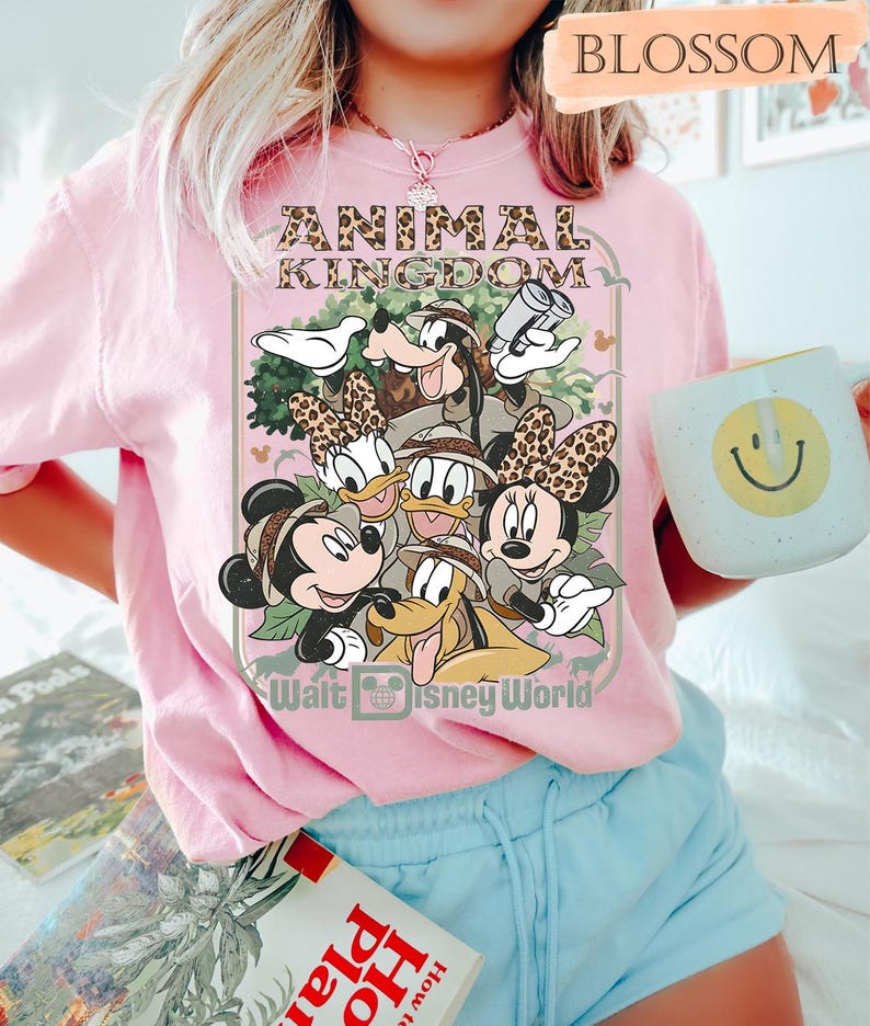May include: Pink t-shirt featuring a graphic of Disney characters, including Mickey, Minnie, Donald, Daisy, Goofy, and Pluto, with the text "ANIMAL KINGDOM" and "Walt Disney World". A white mug with a smiley face is held in the hand.
