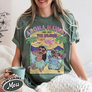 May include: A moss green Comfort Colors t-shirt with a graphic that reads "Aloha Hawaii, Come Visit The Islands." The design showcases cartoon characters in sunglasses and hula skirts, set against a tropical beach scene. The shirt is a green colour.