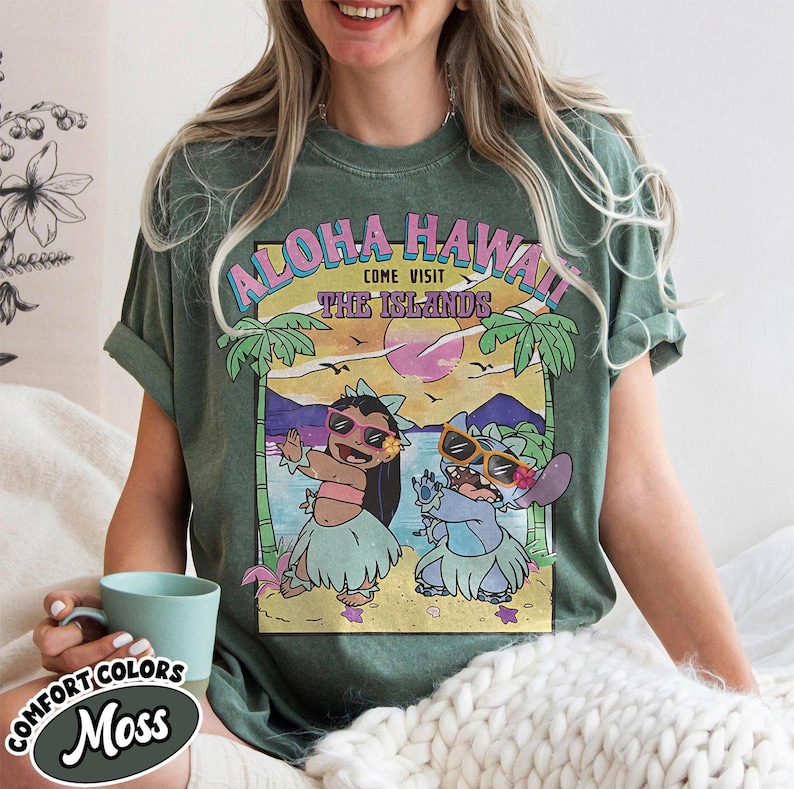 May include: A moss green Comfort Colors t-shirt with a graphic that reads "Aloha Hawaii, Come Visit The Islands." The design showcases cartoon characters in sunglasses and hula skirts, set against a tropical beach scene. The shirt is a green colour.