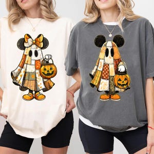 May include: Two t-shirts featuring a cartoon ghost character with Mickey Mouse ears, a patchwork design, and a jack-o'-lantern. One shirt is cream-colored, the other is gray. The ghost is wearing orange shoes and holding a pumpkin.