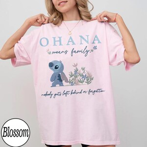 May include: A light pink t-shirt with the text "OHANA means family." and a cartoon character illustration. The illustration features a blue alien-like creature and floral accents. The phrase "nobody gets left behind or forgotten" is also printed on the shirt.