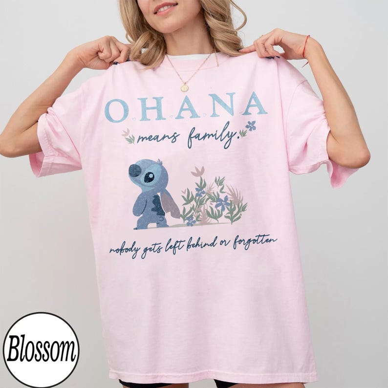 May include: A light pink t-shirt with the text "OHANA means family." and a cartoon character illustration. The illustration features a blue alien-like creature and floral accents. The phrase "nobody gets left behind or forgotten" is also printed on the shirt.