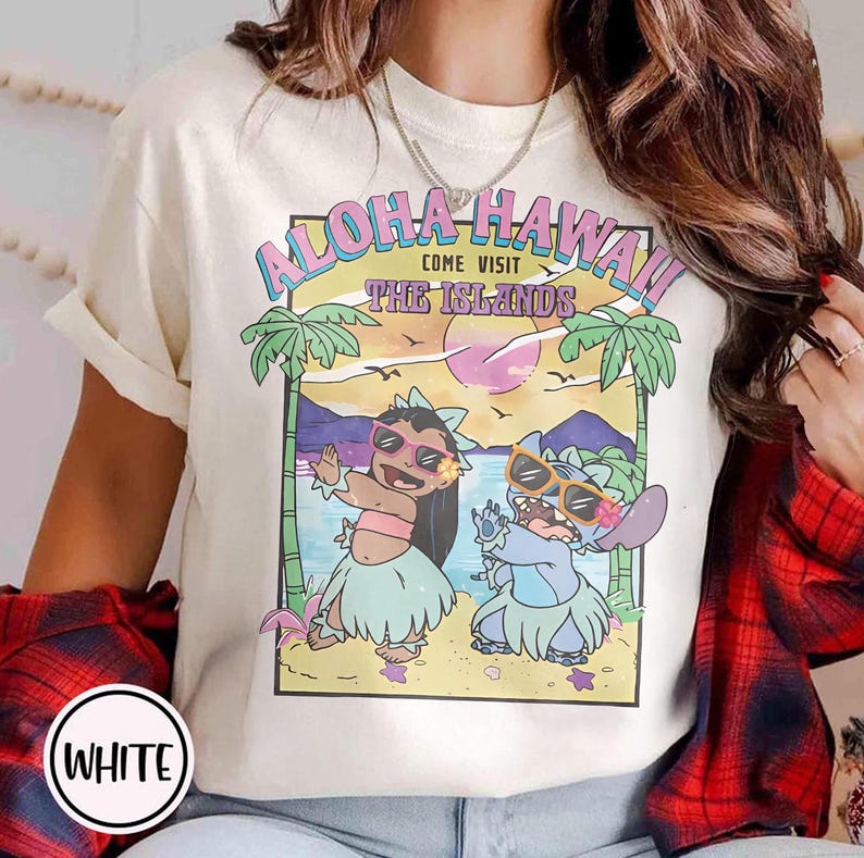 May include: Cream-coloured t-shirt with a vibrant graphic that reads "Aloha Hawaii! Come Visit The Islands." The design showcases cartoon characters, palm trees, and a sunset. The tee has a round neck and short sleeves.