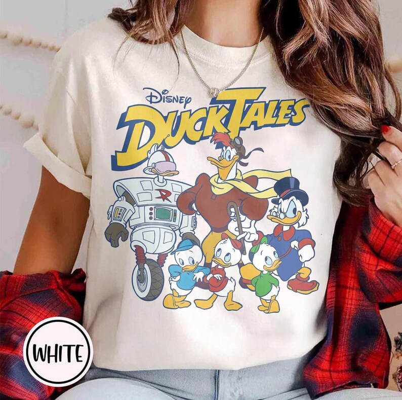May include: Cream-colored t-shirt featuring the Disney DuckTales logo and a graphic of the DuckTales characters. The graphic includes Scrooge McDuck, Huey, Dewey, Louie, Webby, Launchpad McQuack, and Gizmoduck. The shirt has short sleeves.