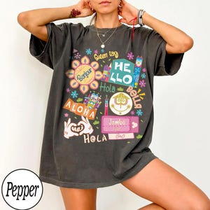 May include: A charcoal gray short-sleeve t-shirt featuring a colorful graphic print with various international greetings including Bonjour, Guten tag, HELLO, Hola, Bella, ALOHA, Jambo, and Hei. The design includes stylized flowers and icons.