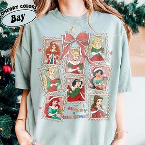 May include: A light green t-shirt featuring a design of various princesses in Christmas-themed postage stamps. A large red bow is at the top, with the words "Merry Christmas" below the stamps. The shirt has the words "Comfort Colors Bay" at the top.