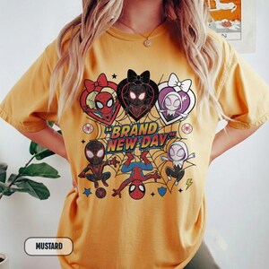 May include: Mustard-colored t-shirt featuring a graphic of Spider-Man characters in heart-shaped designs and the text "BRAND NEW DAY". The design includes various Spider-Man characters and a spider web background.