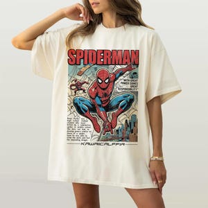 May include: Cream-colored t-shirt featuring a comic book-style illustration of Spider-Man. The design includes the word "SPIDERMAN" in red, and the phrase "WITH GREAT POWER COMES GREAT RESPONSIBILITY". The shirt also has text at the bottom that says "KAWAIICALFFIA".