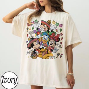 May include: Ivory-coloured t-shirt with a vibrant graphic of Disney characters Mickey Mouse, Minnie Mouse, Donald Duck, Daisy Duck, Goofy, and Pluto, wearing sombreros. The design includes text and decorative elements.