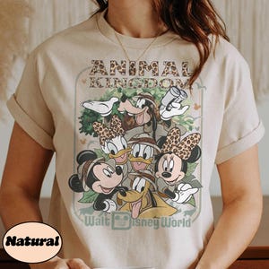 May include: Beige t-shirt featuring a graphic of Disney characters, including Mickey and Minnie Mouse, Goofy, Donald and Daisy Duck, and Pluto. The design includes the text "ANIMAL KINGDOM" and "Walt Disney World".