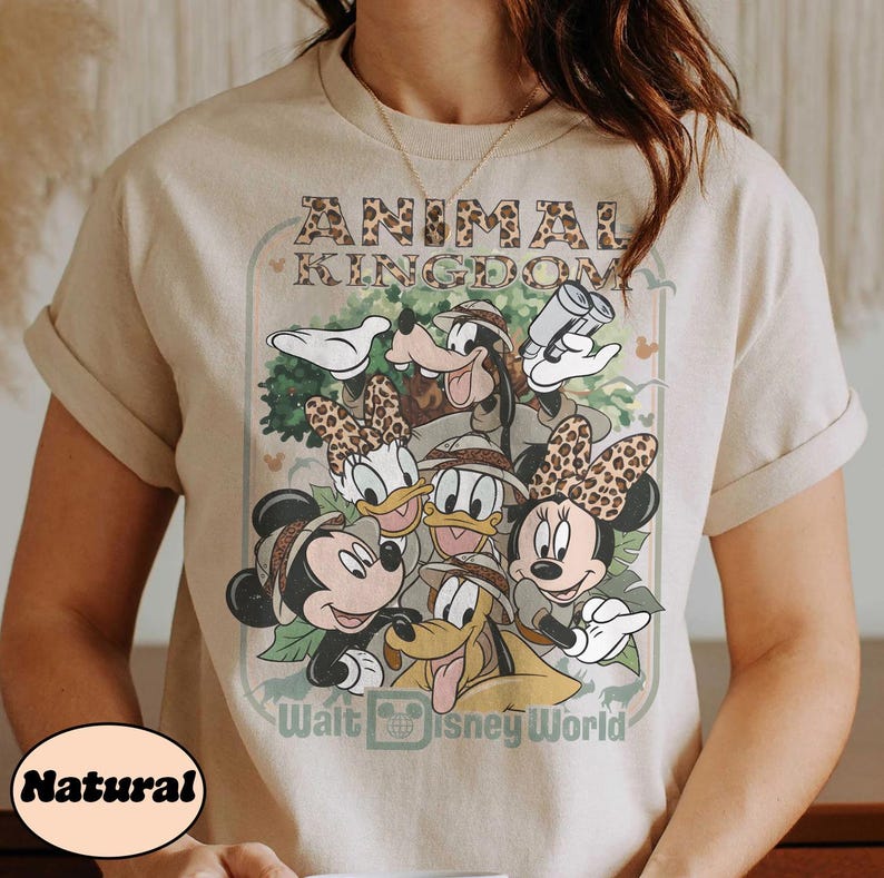 May include: Beige t-shirt featuring a graphic of Disney characters, including Mickey and Minnie Mouse, Goofy, Donald and Daisy Duck, and Pluto. The design includes the text "ANIMAL KINGDOM" and "Walt Disney World".