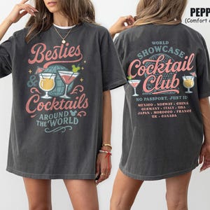 May include: Dark gray t-shirt with the text "Besties & Cocktails Around The World" on the front and "World Showcase Cocktail Club" on the back. The design includes cocktail glasses and a globe. The shirt is a comfort color.