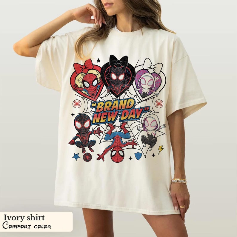 May include: Ivory-colored t-shirt featuring a colorful graphic of Spider-Man characters in heart-shaped designs, with the text "BRAND NEW DAY". The shirt is a Comfort Color brand.