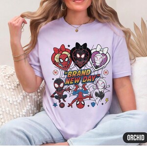 May include: A light purple t-shirt featuring a graphic design with multiple Spider-Man characters in heart-shaped frames. The text "BRAND NEW DAY" is prominently displayed. The shirt has a crew neck and short sleeves.