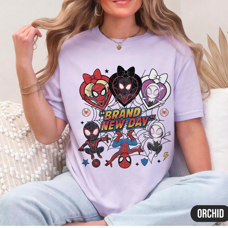 May include: A light purple t-shirt featuring a graphic design with multiple Spider-Man characters in heart-shaped frames. The text "BRAND NEW DAY" is prominently displayed. The shirt has a crew neck and short sleeves.