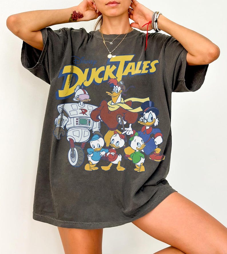 May include: Dark gray t-shirt featuring the Disney DuckTales logo in yellow and a graphic of the DuckTales characters. The shirt has a vintage, worn look. The shirt is a short-sleeved, oversized fit.